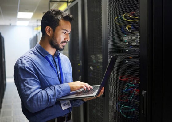 An IT employee with a laptop, with IT equipment in the background 